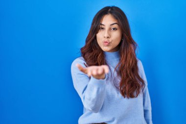 Hispanic young woman standing over blue background looking at the camera blowing a kiss with hand on air being lovely and sexy. love expression. 