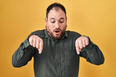 Plus size hispanic man with beard standing over yellow background pointing down with fingers showing advertisement, surprised face and open mouth 