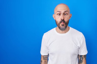 Hispanic man with tattoos standing over blue background making fish face with lips, crazy and comical gesture. funny expression. 