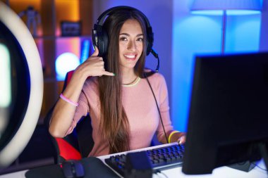 Young hispanic woman playing video games smiling doing phone gesture with hand and fingers like talking on the telephone. communicating concepts. 