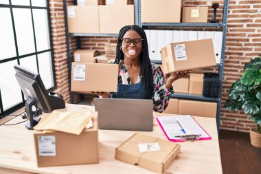 African woman with braids working at small business ecommerce holding packages sticking tongue out happy with funny expression. 
