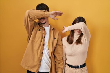 Young hispanic couple standing over yellow background covering eyes with arm, looking serious and sad. sightless, hiding and rejection concept 