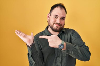 Plus size hispanic man with beard standing over yellow background amazed and smiling to the camera while presenting with hand and pointing with finger. 