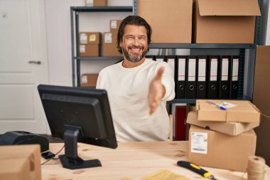 Handsome middle age man working at small business ecommerce smiling friendly offering handshake as greeting and welcoming. successful business. 