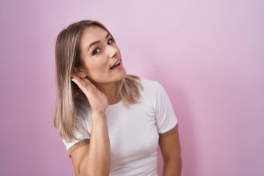 Blonde caucasian woman standing over pink background smiling with hand over ear listening an hearing to rumor or gossip. deafness concept. 