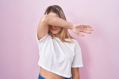 Young blonde woman standing over pink background covering eyes with arm, looking serious and sad. sightless, hiding and rejection concept 