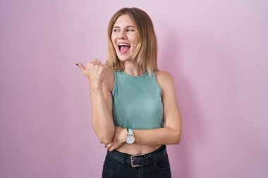 Blonde caucasian woman standing over pink background smiling with happy face looking and pointing to the side with thumb up. 