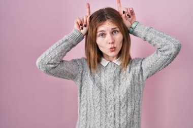 Beautiful woman standing over pink background doing funny gesture with finger over head as bull horns 