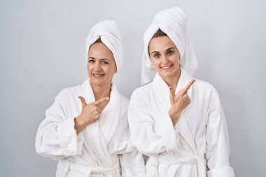 Middle age woman and daughter wearing white bathrobe and towel cheerful with a smile of face pointing with hand and finger up to the side with happy and natural expression on face 