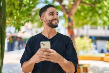 Genç Arap adam parkta akıllı telefon kullanarak kendinden emin gülümsüyor.