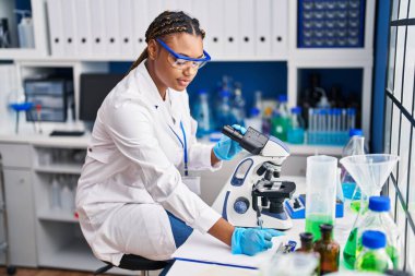 African american woman scientist using microscope writing document at laboratory