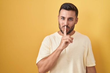 Handsome hispanic man standing over yellow background asking to be quiet with finger on lips. silence and secret concept. 
