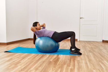 Spor merkezinde formda top kullanan genç Latin kadın karın kası egzersizi yapıyor.