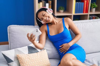 Genç, güzel, Latin bir kadın akıllı telefondan video izliyor. Evdeki kanepede oturuyor.