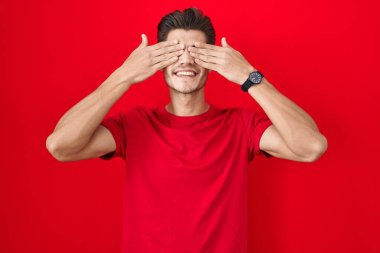 Young hispanic man standing over red background covering eyes with hands smiling cheerful and funny. blind concept. 