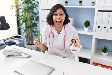 Young hispanic doctor woman holding anatomical model of uterus with fetus and birth control pills celebrating crazy and amazed for success with open eyes screaming excited. 
