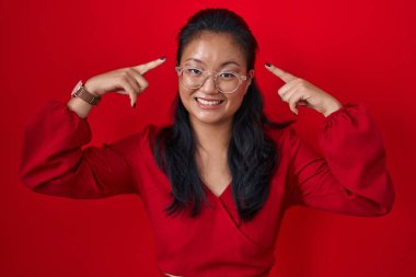 Asian young woman standing over red background smiling pointing to head with both hands finger, great idea or thought, good memory 