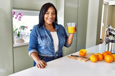 Hispanic brunette woman drinking a glass of fresh orange juice at the kitchen