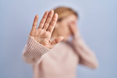 Mavi arka planda dikilen İspanyol kadın elleriyle gözlerini kapatıyor ve üzgün ve korku ifadeleriyle jest yapıyor. utangaç ve olumsuz kavram. 