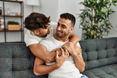 İki İspanyol erkek, kendilerine güvenerek gülümsüyor ve evde birbirlerine sarılıyorlar.