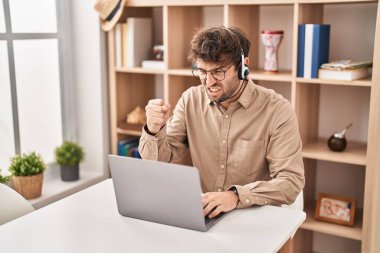 Hispanic young man wearing call center agent headset annoyed and frustrated shouting with anger, yelling crazy with anger and hand raised 