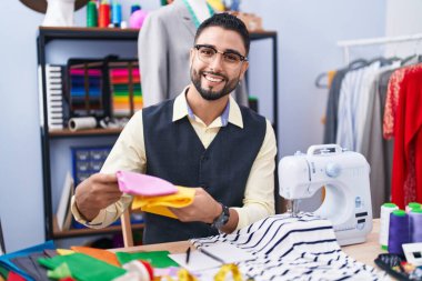 Young arab man tailor smiling confident holding cloths at clothing factory