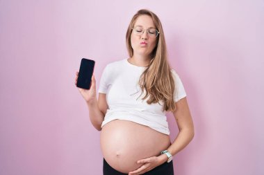 Young pregnant woman showing belly holding smartphone looking at the camera blowing a kiss being lovely and sexy. love expression. 