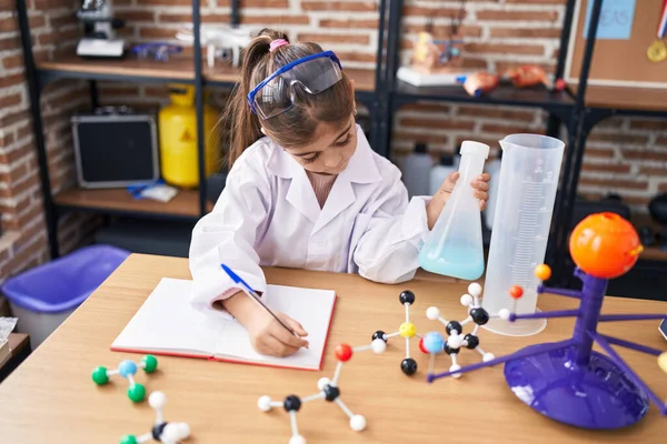 Adorable hispanic girl student holding test tube writing on notebook at laboratory classroom