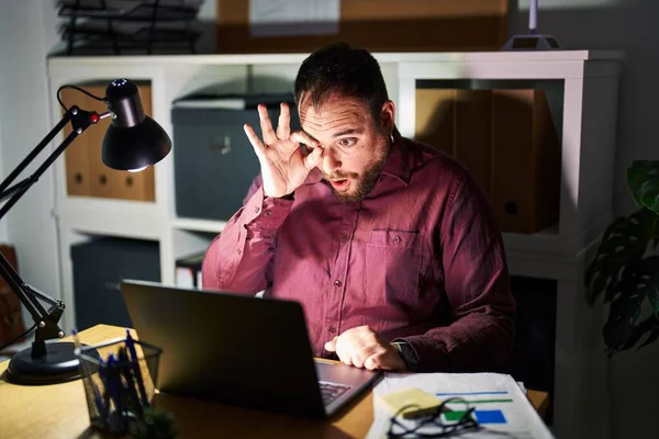 Plus size hispanic man with beard working at the office at night doing ok gesture shocked with surprised face, eye looking through fingers. unbelieving expression.