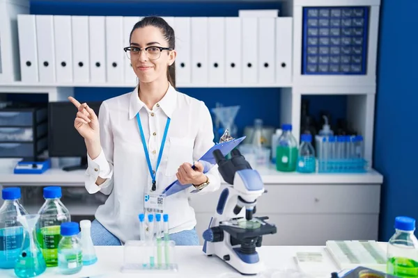 Young brunette woman working at scientist laboratory pointing with hand finger to the side showing advertisement, serious and calm face