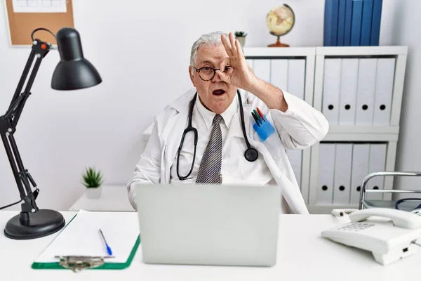 Senior caucasian man wearing doctor uniform and stethoscope at the clinic doing ok gesture shocked with surprised face, eye looking through fingers. unbelieving expression.