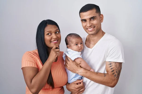 Young hispanic couple with baby standing together over isolated background with hand on chin thinking about question, pensive expression. smiling and thoughtful face. doubt concept.
