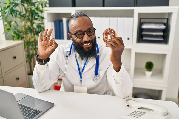 Hombre Afroamericano Que Trabaja Clínica Dietista Sosteniendo Donut Haciendo Signo —  Fotos de Stock