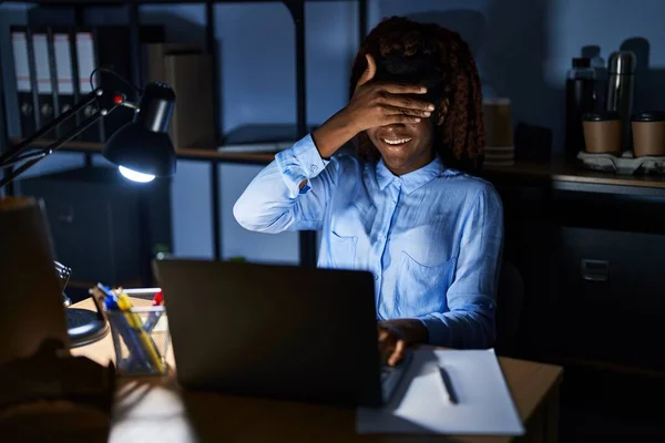 African woman working at the office at night smiling and laughing with hand on face covering eyes for surprise. blind concept.