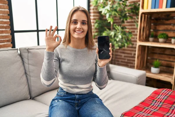 Young woman holding broken smartphone showing cracked screen doing ok sign with fingers, smiling friendly gesturing excellent symbol