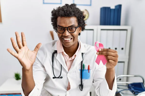 African doctor man holding pink cancer ribbon celebrating victory with happy smile and winner expression with raised hands