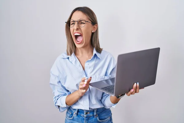 Young woman working using computer laptop angry and mad screaming frustrated and furious, shouting with anger. rage and aggressive concept.