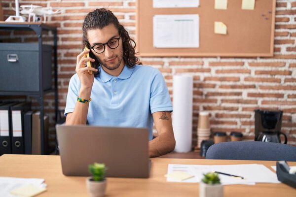Young hispanic man business worker using laptop talking on smartphone at office