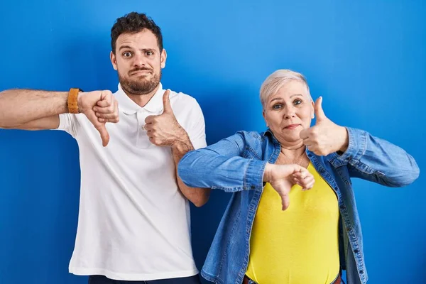 青い背景の上に立つ若いブラジル人の母親と息子は 親指を上下に 意見の相違や合意表現をしています 狂気の葛藤 — ストック写真
