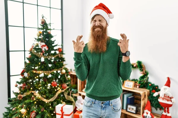 Redhead man with long beard wearing christmas hat by christmas tree gesturing finger crossed smiling with hope and eyes closed. luck and superstitious concept.