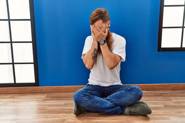 Redhead man with long beard sitting on the floor at empty room with sad expression covering face with hands while crying. depression concept.
