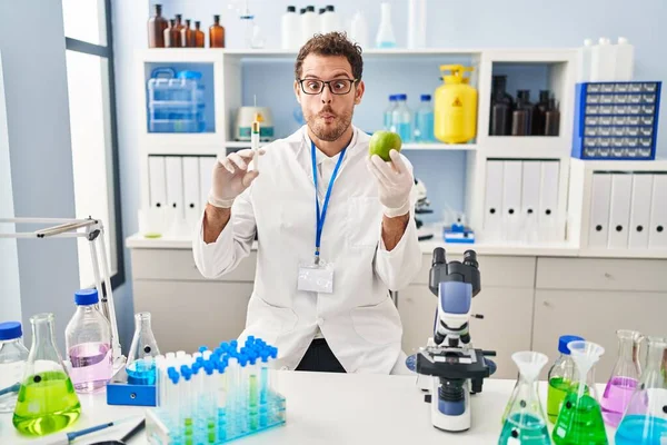 Young hispanic man working at scientist laboratory holding apple making fish face with mouth and squinting eyes, crazy and comical.