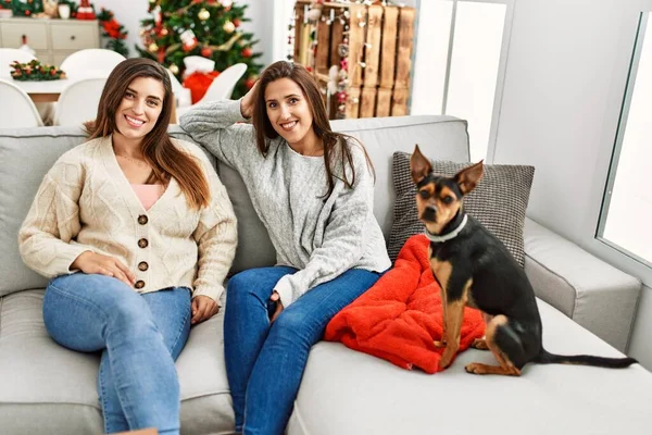 Two women smiling confident sitting with dog by christmas tree at home
