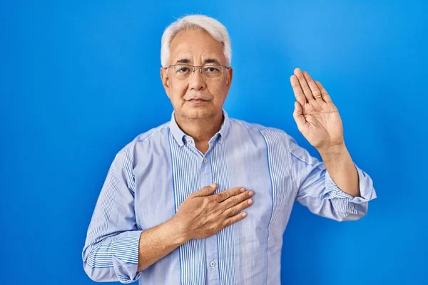 Hispanic senior man wearing glasses swearing with hand on chest and open palm, making a loyalty promise oath