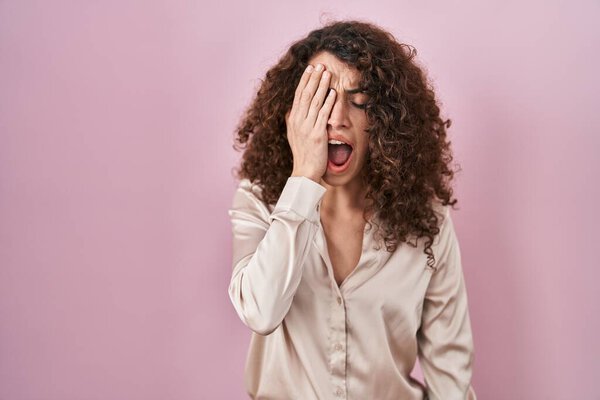Латиноамериканка с вьющимися волосами, стоящими на розовом фоне, зевая усталая, прикрывающая рукой половину лица, глаза и рот. лицо болит от боли. 