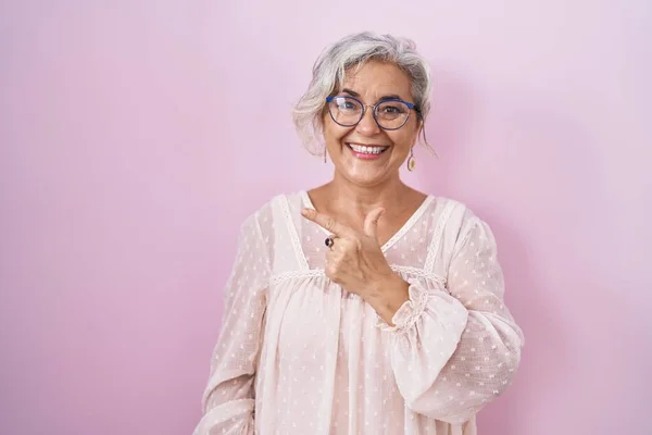 Middle age woman with grey hair standing over pink background cheerful with a smile of face pointing with hand and finger up to the side with happy and natural expression on face
