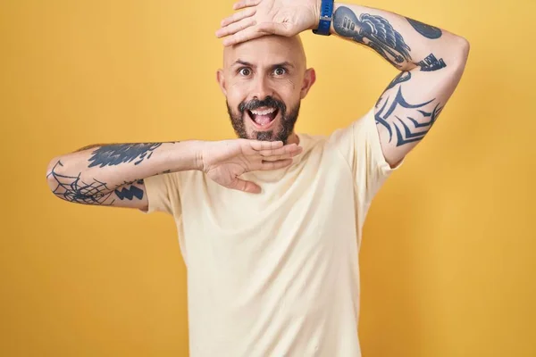 Hispanic man with tattoos standing over yellow background smiling cheerful playing peek a boo with hands showing face. surprised and exited