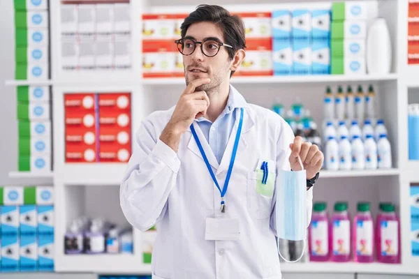 Young Hispanic Man Working Pharmacy Drugstore Holding Safety Mask Serious — Stockfoto