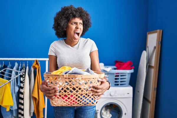 Black woman with curly hair holding laundry basket angry and mad screaming frustrated and furious, shouting with anger. rage and aggressive concept.