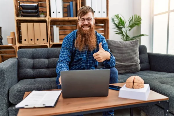 Redhead man with long beard using laptop working at psychology clinic smiling happy and positive, thumb up doing excellent and approval sign